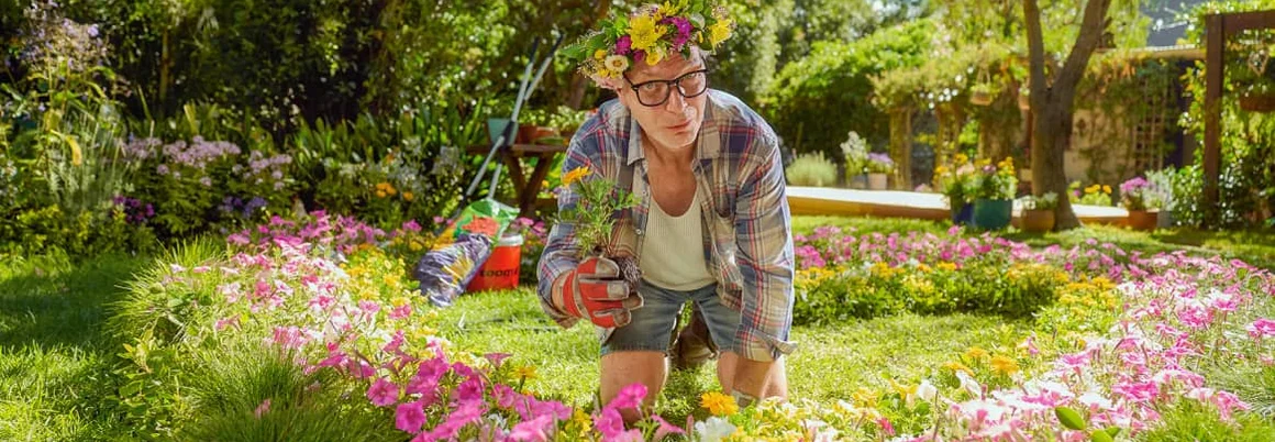 Eine Person mit Blumenkranz auf dem Kopf kniet im Garten zwischen Blumen.